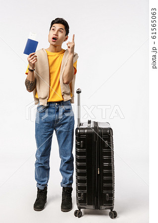 Full-length vertical shot asian male model 20s in yellow t-shirt, jeans, raise index finger eureka gesture, have great idea, came up with where travel, holding passport, standing airport with luggage 61565063