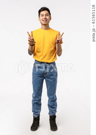 Vertical full-length shot charming chinese guy in yellow t-shirt, jeans, showing peace sign and smiling happy, express positivity, having fun, standing entertained and joyful, white background 61565116