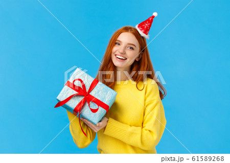Holidays, celebration and presents concept. Cheerful redhead girl love christmas, enjoy celebrating with friends, receive gift in wrapped box from secret santa, wearing cute hat, blue background 61589268