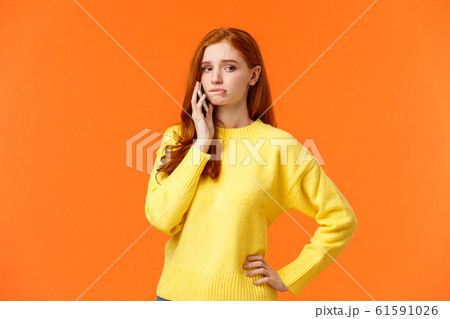 Girl catching anxiety calling someone. Perplexed or nervous cute redhead woman biting lip and looking away as having conversation on mobile phone, standing orange background sad 61591026