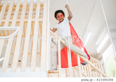 Adorable boy in mantle of superman holding by rope while gliding down railings Adorable boy in mantle of superman holding by rope while gliding down railings 61599399