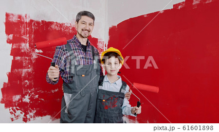 A boy and his smiling father standing in the room and holding rollers covered in red painting A boy and his smiling father standing in the room and holding rollers covered in red painting 61601898