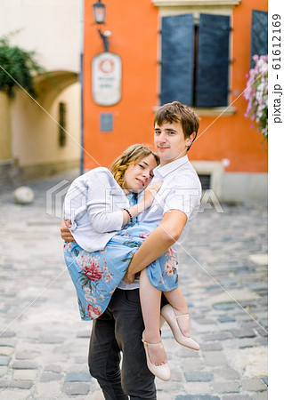 smiling young handsome man holding on hands his happy disabled girlfriend, while standing outdoors, on the background of old orange building smiling young handsome man holding on hands his happy disabled girlfriend, while standing outdoors, on the background of old orange building 61612169