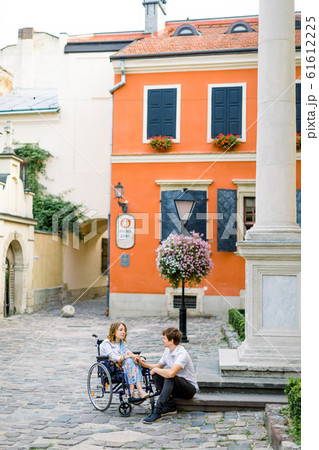 Pretty young smiling woman in the wheelchair and handsome man sitting on the stone stairs, looking each other in love, walking in the old city center. Bright orange building on the background Pretty young smiling woman in the wheelchair and handsome man sitting on the stone stairs, looking each other in love, walking in the old city center. Bright orange building on the background 61612225