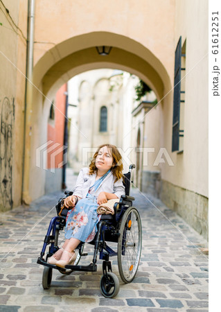 Portrait of young blond-haired woman sitting in wheelchair and looking at camera, walking outdoors in old city center, building arch on the background 61612251
