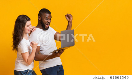 Happy interracial spouses celebrating win with laptop, raising fists in excitement 61661549