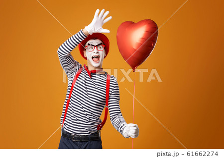 Happy mime male in red hat and vest holds heart ball in his hands on empty orange background 61662274
