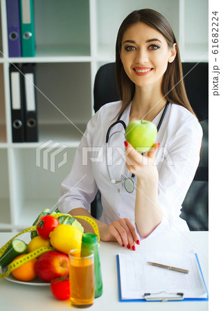 Health. Diet and Healthy Nutrition. Doctor Dietitian Holding in Hands Fresh Green Apple and Centimeter Ribbon. Smiles Beautiful and Young Doctor in the Light Room. High Resolution Health. Diet and Healthy Nutrition. Doctor Dietitian Holding in Hands Fresh Green Apple and Centimeter Ribbon. Smiles Beautiful and Young Doctor in the Light Room. High Resolution 61682224