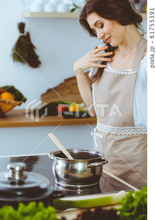 Young brunette woman cooking soup in kitchen. Housewife holding wooden spoon in her hand. Food and health concept Young brunette woman cooking soup in kitchen. Housewife holding wooden spoon in her hand. Food and health concept 61755391