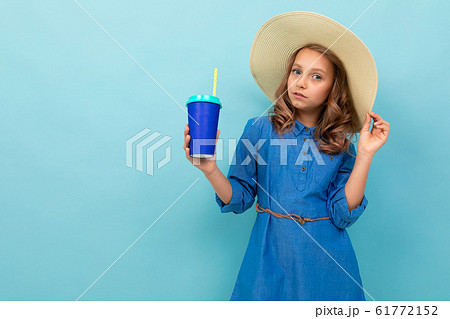 Caucasian girl with wavy brown hair in blue dress, big hat holds a cup of juice isolated on blue Caucasian girl with wavy brown hair in blue dress, big hat holds a cup of juice isolated on blue 61772152