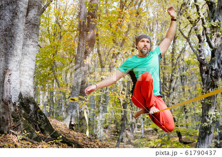 Wide angle male tightrope walker balancing while sitting barefoot on slackline in autumn forest. The concept of outdoor sports and active life of people aged 61790437