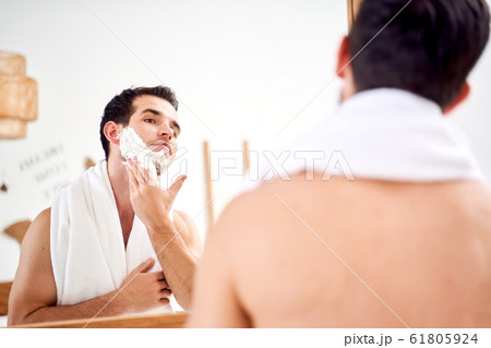 Brunet man with towel on shoulders spreads face with shaving foam while standing in bath near mirror 61805924