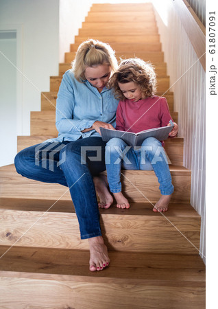 A cute small girl with mother indoors at home, reading on staircase. A cute small girl with mother indoors at home, reading on staircase. 61807091