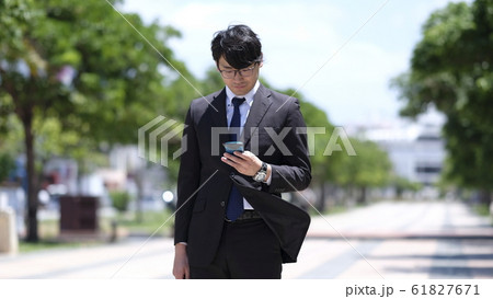晴れた朝の駅前で出勤途中にスマホを見るスーツ姿の眼鏡をかけた20代の日本人の男性 晴れた朝の駅前で出勤途中にスマホを見るスーツ姿の眼鏡をかけた20代の日本人の男性 61827671