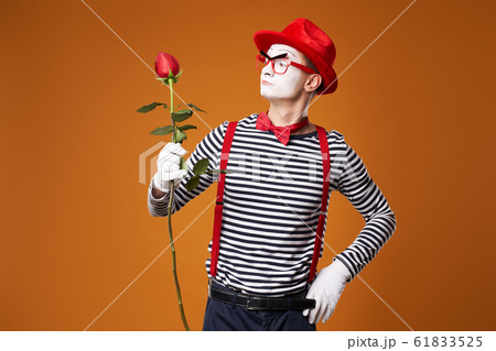 Serious mime in red hat, glasses and vest with rose in hands on orange background Serious mime in red hat, glasses and vest with rose in hands on orange background 61833525