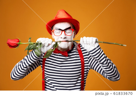 Close up mime with white face in red hat and striped T-shirt holding rose in his mouth on blank orange background 61833670