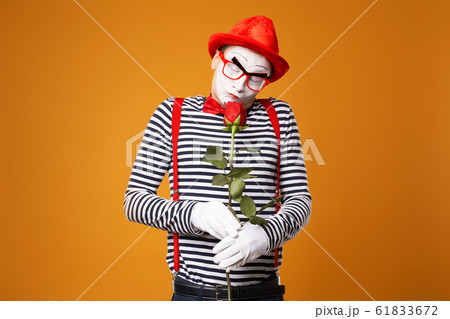 Pensive mime man with white face in red hat and striped t-shirt holding rose isolated on empty orange background 61833672