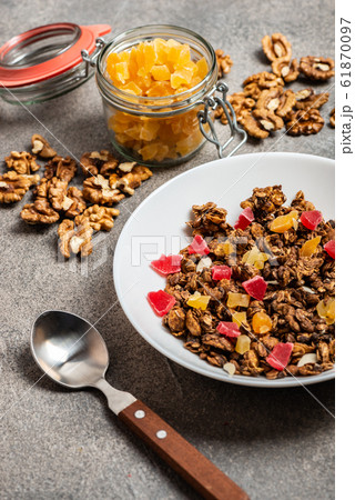 Granola with dried fruits in a white plate on a gray background. Close-up. Open decorative jar with dried fruits. 61870097