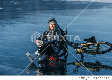 Woman sitting near her bike on ice. The girl cyclist stopped to rest. She sits on the wheel and enjoys the beautiful view of the sunset. Woman sitting near her bike on ice. The girl cyclist stopped to rest. She sits on the wheel and enjoys the beautiful view of the sunset. 61877732