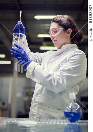Young lab woman in glasses and white coat with experimental glass in her hands conducts experiments 61892907