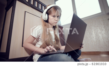 A smart little girl of seven years old in white headphones with a laptop in her hands is pushing on the floor in her room. The young generation on the Internet and IT technology 61901010