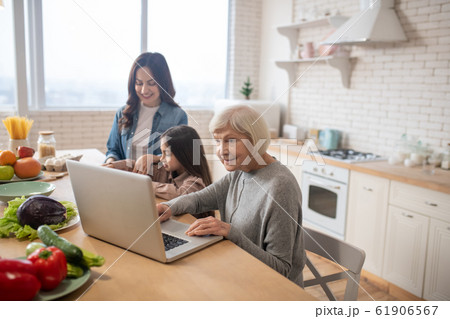 Grandmother with laptop and mom with daughter making breakfast. Grandmother with laptop and mom with daughter making breakfast. 61906567