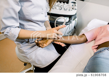 Cropped image of hands of female doctor with ultrasound scanner conducting ultrasound examination of wrist of young African woman in clinic Cropped image of hands of female doctor with ultrasound scanner conducting ultrasound examination of wrist of young African woman in clinic 61908708