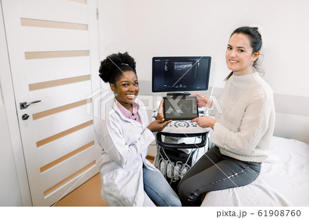 Happy Caucasian pregnant woman visit doctor at hospital or medical clinic for pregnancy consultant. African woman gynecologist with patient holds the digital tablet with photo of the baby and smiling 61908760