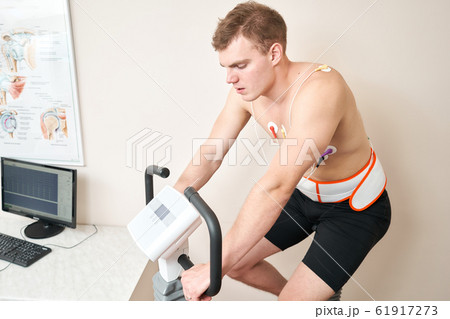 Man patient, pedaling on a bicycle ergometer stress test system for the function of heart checked. Athlete does a cardiac stress test in a medical study, monitored by the doctor. 61917273