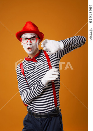 Serious young mime in white gloves and red hat looking at camera on orange background 61918964