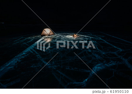 Fire on ice at night. Campground on ice. Tent stands next to bonfire. Lake Baikal. Nearby there is car. Shelter tent and ice are illuminated from the inside. Beautiful bonfire on real cracked ice. 61950110