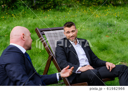Businessmen are sitting in chairs and talking on grass outdoors 61982966
