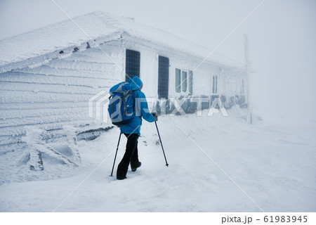 Hiker in winterwear using trekking sticks while passing by snowy building Hiker in winterwear using trekking sticks while passing by snowy building 61983945