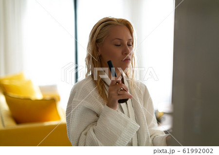 Young beautiful model in a waffle bathrobe gown with closed eyes and a make-up brush in her hands is standing in an apartment with yellow furniture. Young beautiful model in a waffle bathrobe gown with closed eyes and a make-up brush in her hands is standing in an apartment with yellow furniture. 61992002