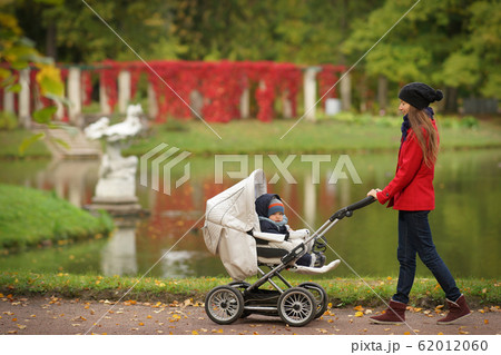 Young woman in red coat walking in park with little child in baby carriage, background of colorful autumn leaves Young woman in red coat walking in park with little child in baby carriage, background of colorful autumn leaves 62012060