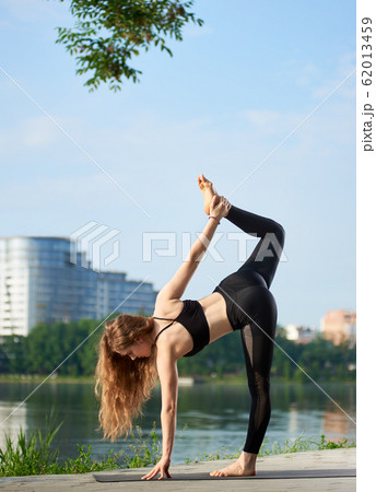 Flexible yoga female with long hair standing in sugarcane pose, ardha chandra chapasana near the river at city park 62013459