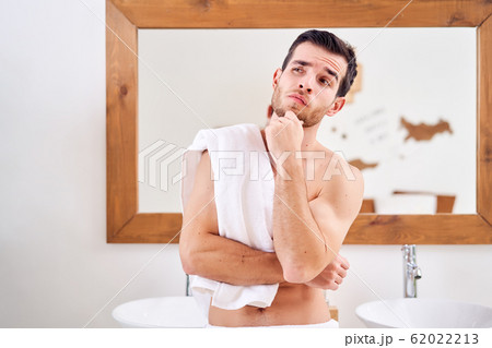Pensive brunet male with towel on shoulder stands near mirror in bath Pensive brunet male with towel on shoulder stands near mirror in bath 62022213