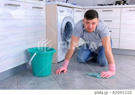 Unhappy tired man in rubber gloves washes the floor in the kitchen and looks at the camera at the end. 62041110