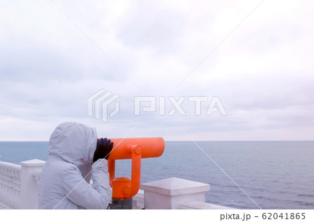 Woman in white jacket and hood looks in binoculars at sea waterfront with sea view. Side view. Woman in white jacket and hood looks in binoculars at sea waterfront with sea view. Side view. 62041865
