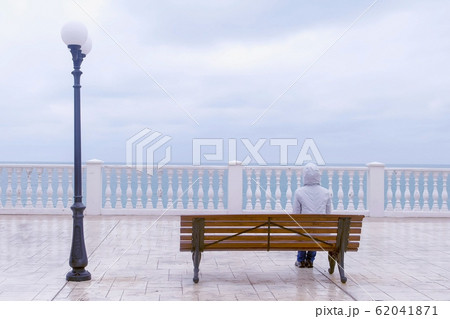 Woman in white jacket and hood is sitting on the bench on beautiful terrace with sea view on waterfront. Back view. Woman in white jacket and hood is sitting on the bench on beautiful terrace with sea view on waterfront. Back view. 62041871