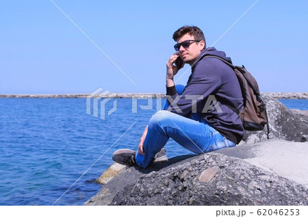 Man traveller calls smartphone sits on stone waits answer on sea background. 62046253