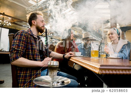 Close-up of Young man in casual wear smoking traditional hookah pipe. man exhaling smoke. Friends in cafe having fun 62058603