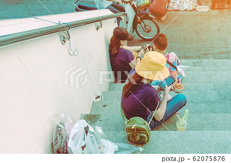 A woman wearing a yellow hat sit to play a smartphone while the other hand hold a cup of meat on the stair in the city A woman wearing a yellow hat sit to play a smartphone while the other hand hold a cup of meat on the stair in the city 62075876