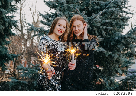 Two young women in evening dress holding sparklers in hand on fir tree background outdoor. Closeup of two beautiful girl holding holding bengal lights at party Two young women in evening dress holding sparklers in hand on fir tree background outdoor. Closeup of two beautiful girl holding holding bengal lights at party 62082598