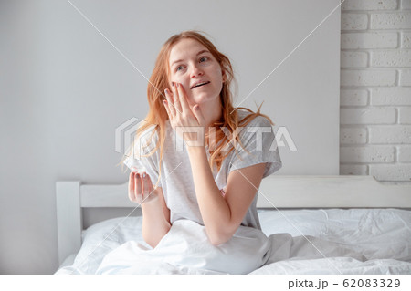 Woman holding bottle of cream with cosmetic products. Blonde woman holding a glass jar with face cream sitting on the bed Woman holding bottle of cream with cosmetic products. Blonde woman holding a glass jar with face cream sitting on the bed 62083329
