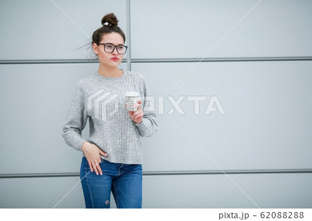 A woman with glasses drinking coffee against a gray wall 62088288