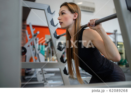 Young fit woman doing squats in gym with training bar, side view. Sport and healthy lifestyle. Young fit woman doing squats in gym with training bar, side view. Sport and healthy lifestyle. 62089268