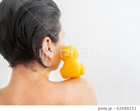 Naked woman with short hair takes a shower. Yellow rubber duck is sitting on her shoulder. Girl with funny toy in white bathroom. Naked woman with short hair takes a shower. Yellow rubber duck is sitting on her shoulder. Girl with funny toy in white bathroom. 62090251