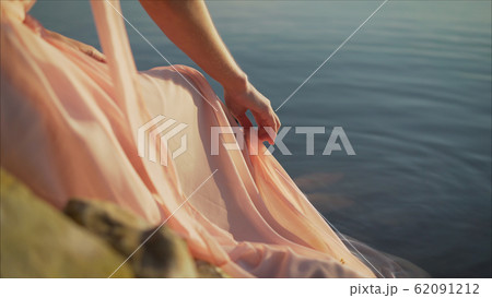 girl in pale pink dress with red hair and backpack walking along river bank, sitting on the sandy shore. The girl in a pink dress on the river bank. 62091212