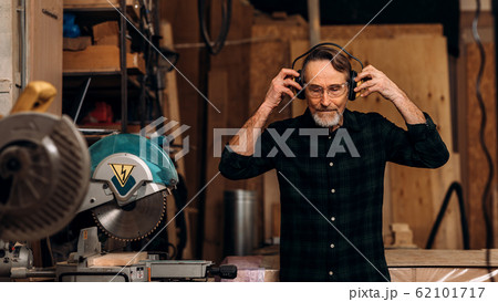 Senior carpenter wearing noise-canceling headphones preparing for working on saw machine Senior carpenter wearing noise-canceling headphones preparing for working on saw machine 62101717